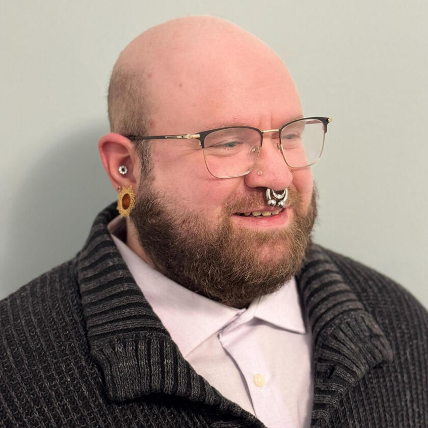 Avi, a white person with a shaved head, beard, glasses, and large silver facial jewelry, smiles and looks to the side.