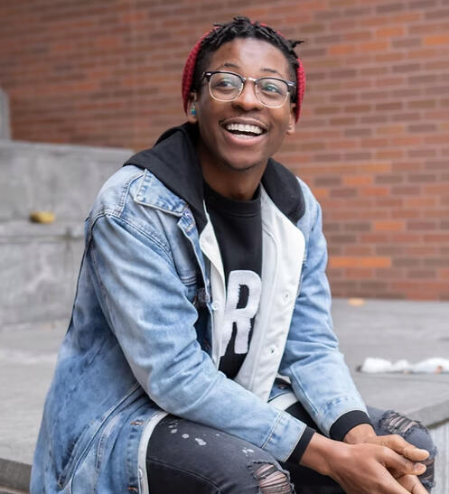 TJ, a black man wearing a denim jacket and glasses, looks to one side of the camera with a wide smile.